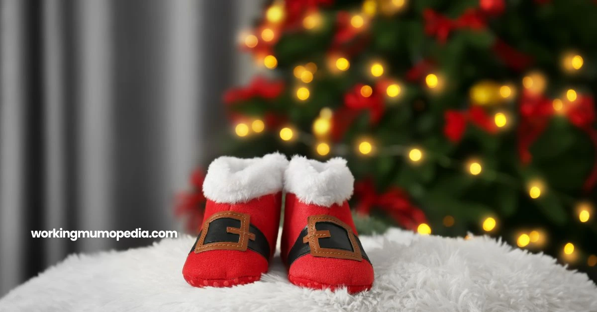 A pair of red baby booties with white fur trim and black belt detailing, resembling Santa's boots, placed on a white fluffy surface. In the background, a beautifully lit Christmas tree with ornaments and warm golden lights adds a festive atmosphere. The text "workingmumopedia.com" is visible on the image.
