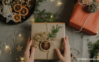 Hands holding a wrapped gift with dried oranges and greenery, surrounded by holiday decorations and twinkling lights on a table.