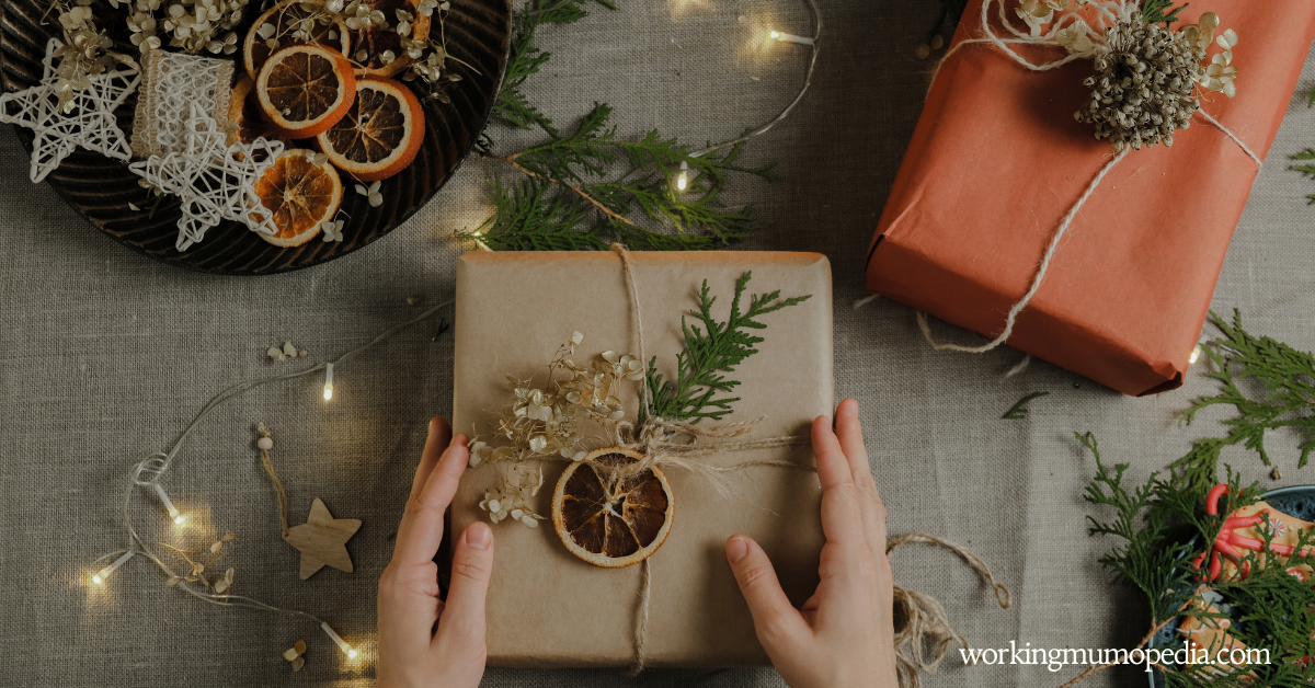 Hands holding a wrapped gift with dried oranges and greenery, surrounded by holiday decorations and twinkling lights on a table.